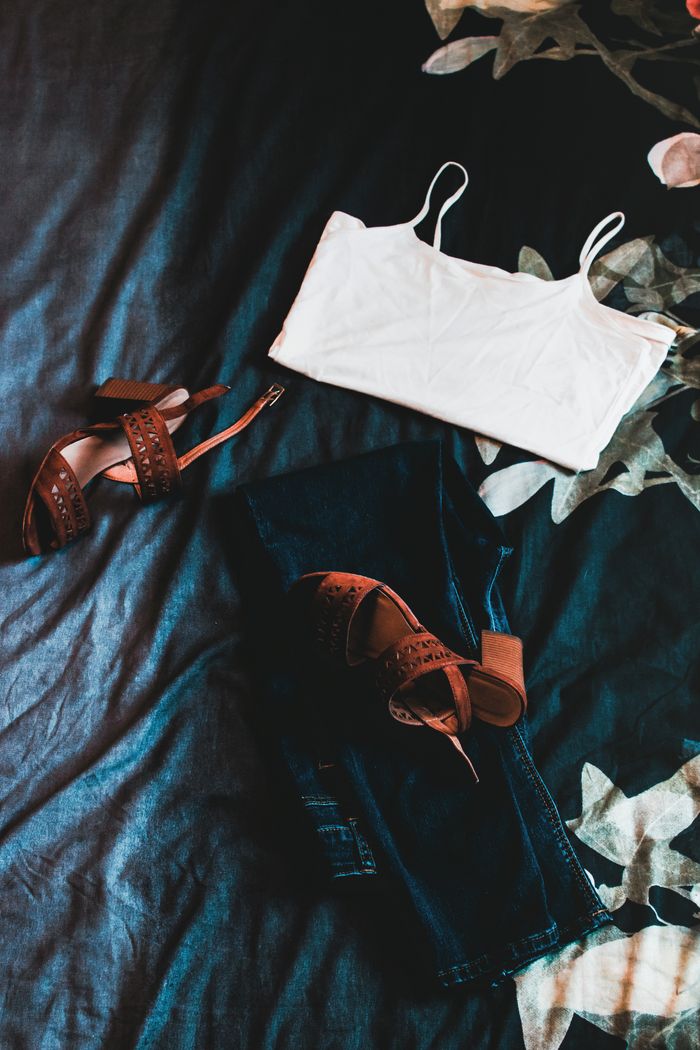 Flat lay of casual outfit with white tank top, jeans, and sandals on floral bedspread.